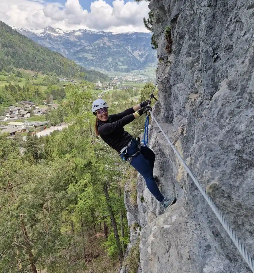 Ella Neff klettert mit Helm an einem Klettersteig an einer Felswand über einem Alpental
