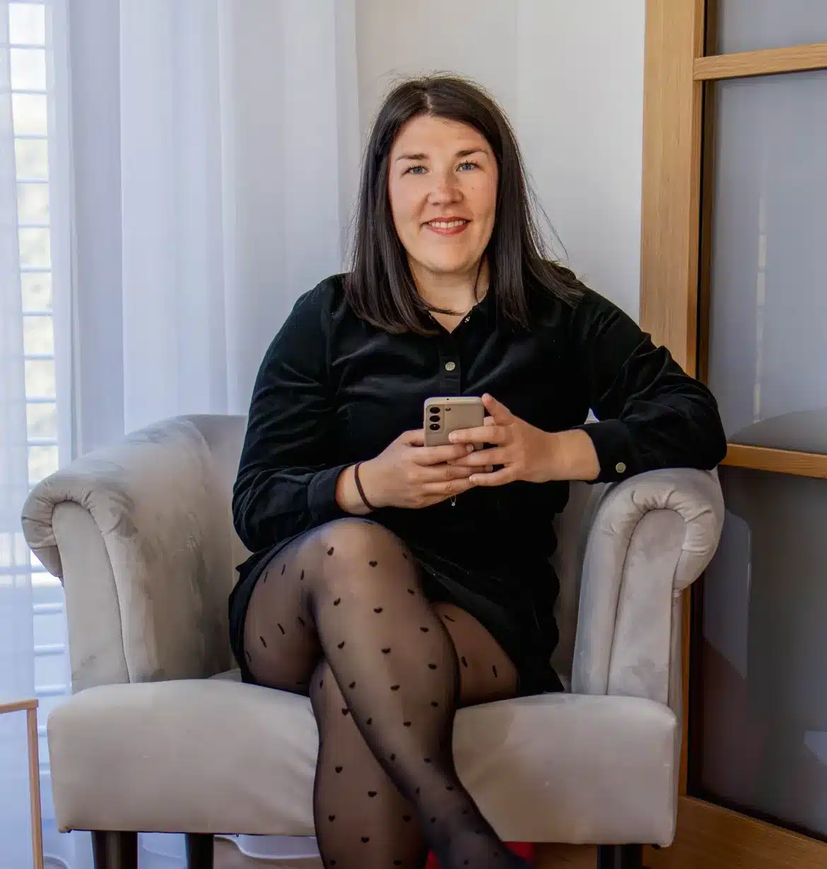 Ella Neff sitzt lächelnd in einem hellen Sessel und hält ein Smartphone in den Händen