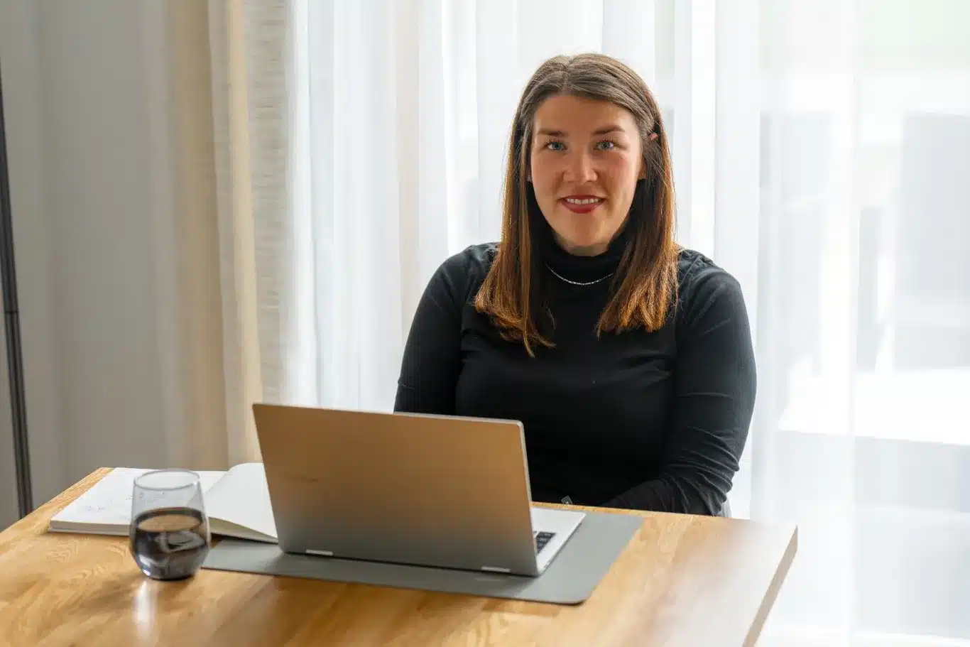 Ella Neff sitzt lächelnd an einem Holztisch mit Laptop und Wasserglas